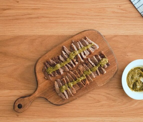 sliced pan-seared steaks on a cutting board with pesto sauce in a bowl with garlic and spices on the side