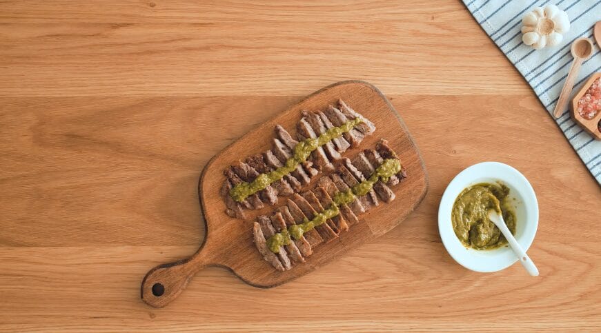 sliced pan-seared steaks on a cutting board with pesto sauce in a bowl with garlic and spices on the side