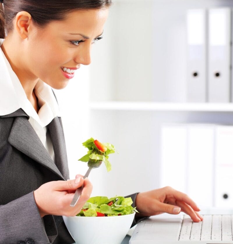 business woman eating salad on her desk while working on laptop