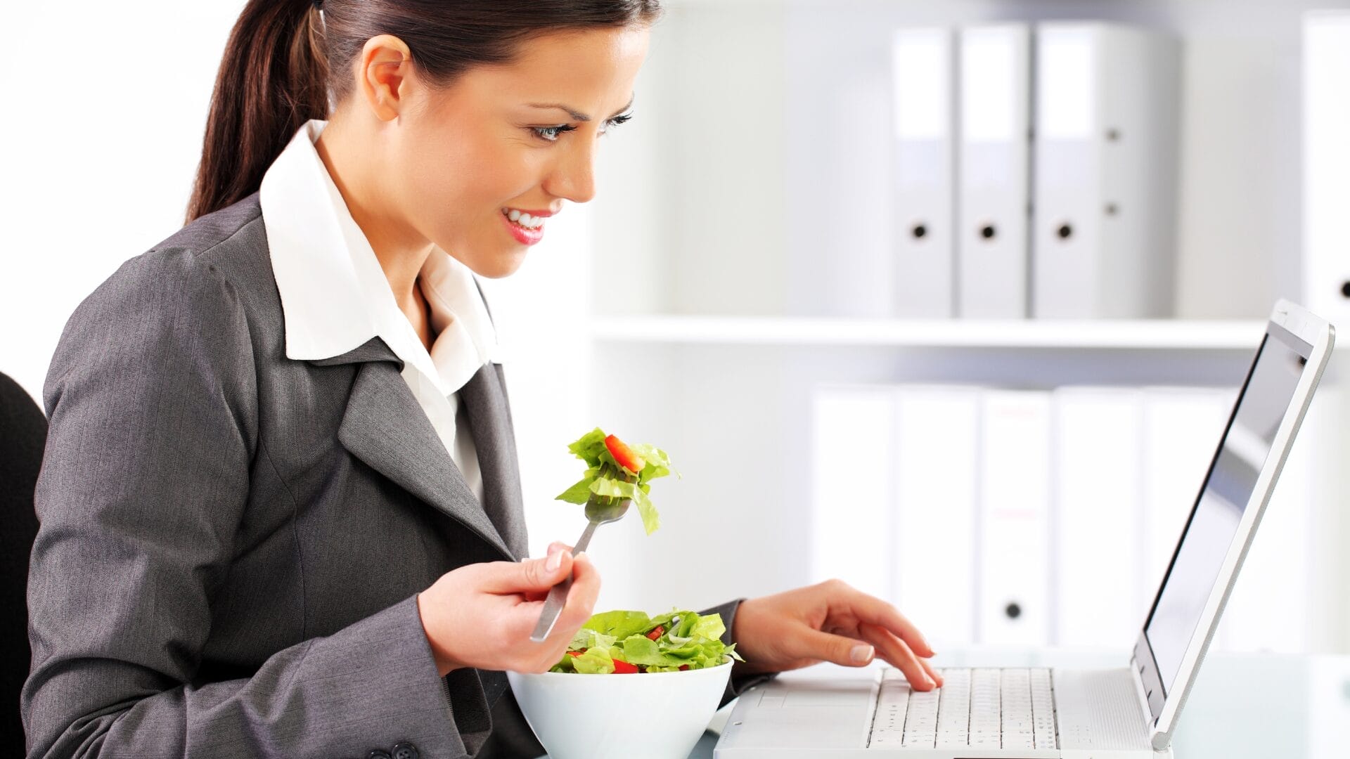 business woman eating salad on her desk while working on laptop