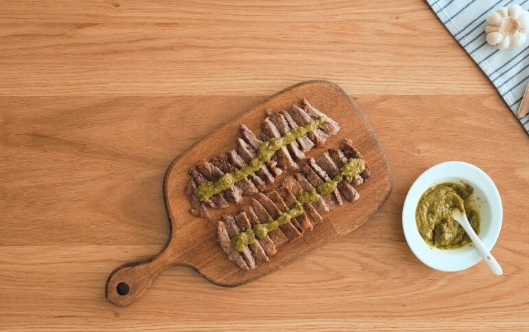 sliced pan-seared steaks on a cutting board with pesto sauce in a bowl with garlic and spices on the side